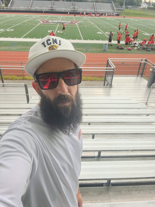 A bearded man wearing a white 'TCNJ' cap, red-tinted sunglasses, and a white shirt takes a selfie in front of a football field and running track bleachers.