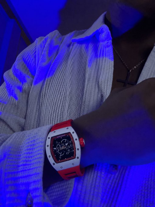 A close-up photo of a person wearing a white ribbed shirt and a cross necklace, showing a red and white Richard Mille-style luxury watch on their wrist under blue lighting.