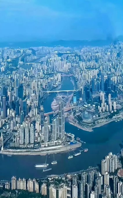 An aerial view of a dense cityscape featuring a river winding through skyscrapers, with bridges crossing the water and boats visible below.