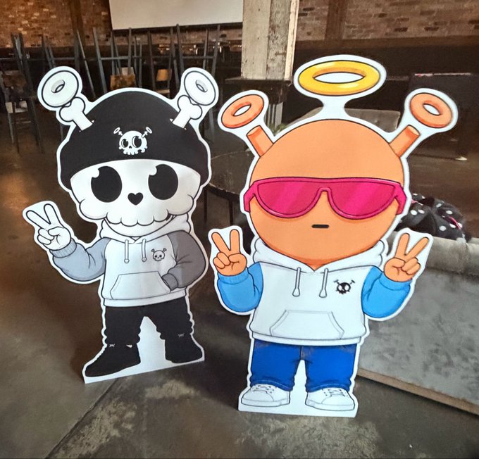 Two cartoon character standees—a skull wearing a beanie and an orange alien with pink sunglasses and a halo, both flashing peace signs in matching hoodies—sit on a couch, representing the creator "papii" being recognized and rewarded for their authentic dedication rather than for clout or extraction. The image illustrates the post's theme of rewarding genuine passion and consistency in the community.