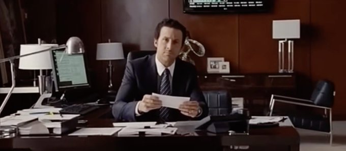 A man in a suit sits at a desk in a corporate office holding a piece of paper, appearing to be from a TV show or film.