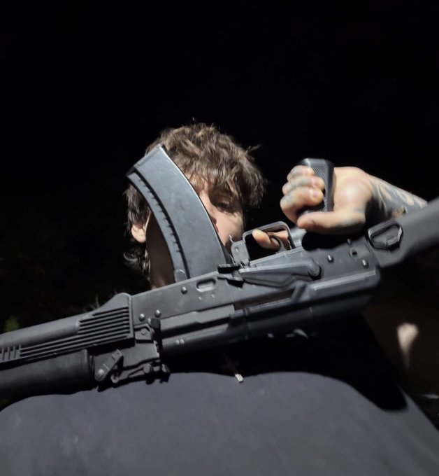 A young person with dark, messy hair grips a black assault rifle with an extended magazine, photographed from a dramatic low angle against a black background. The menacing, action-oriented aesthetic evokes the gritty, heavy metal "gmonad" vibe of the accompanying post.