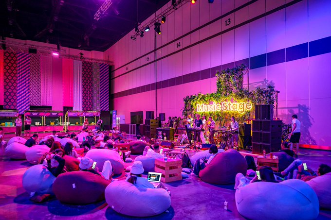A large indoor venue is bathed in pink and purple lighting, featuring a 'MusicStage' sign and a crowd of people relaxing on bean bag chairs while listening to a live performance.