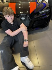 A young man in black clothing crouching next to a race car with orange stripes on a tiled floor.