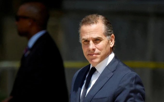 Hunter Biden walks outdoors wearing a dark suit, with a security guard visible in the background.