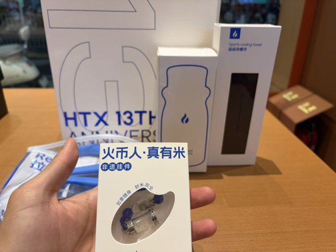 A hand holds a small gift box labeled '火币人·真有米' (HTX 13th Anniversary) containing a USB drive, with an HTX 13th Anniversary water bottle and sports cooling towel visible in the background.