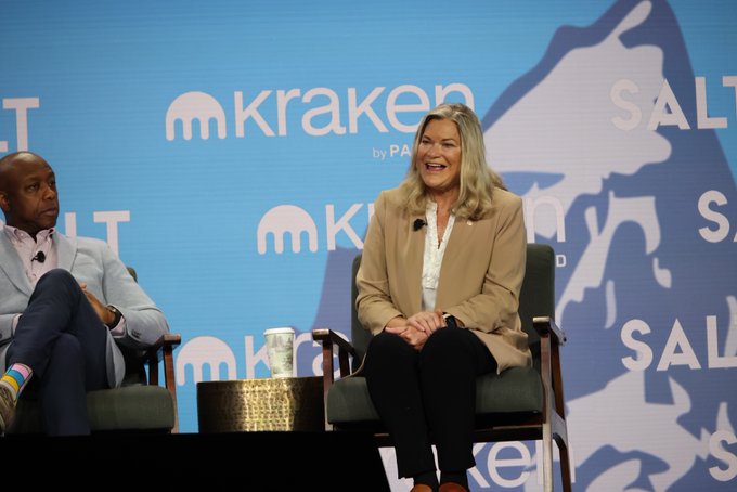 A blonde woman in a tan blazer laughs while seated on stage at the SALT Conference, next to a man in a dark suit, with "Kraken" and "SALT" branding visible on the backdrop behind them.