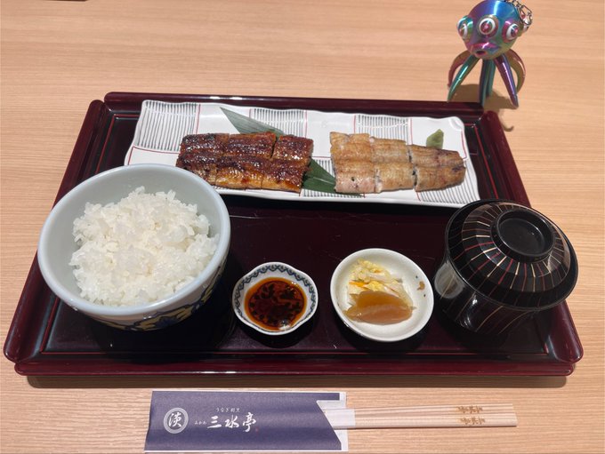 A Japanese teishoku set meal on a dark tray, featuring a bowl of white rice, grilled eel, a side dish of pickled ginger, dipping sauce, miso soup, and chopsticks with the name "三水亭" on the wrapper, next to a small colorful wind chime.
