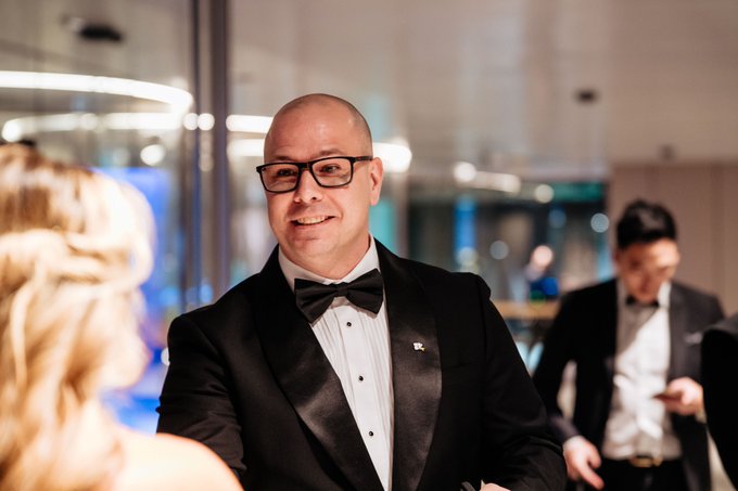 Photograph of a smiling bald man wearing glasses and a black tuxedo with bow tie at what appears to be a formal indoor event, with another similarly dressed guest visible in the background.