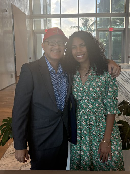A smiling man wearing a red baseball cap, glasses, and a dark blazer over a checkered shirt poses with his arm around a woman with curly hair wearing a green floral dress, as they stand together in a bright lobby with large windows. The post tagging @TheBTCQueen suggests the woman is a notable figure in the Bitcoin community, with the image likely capturing a friendly meeting between the two.