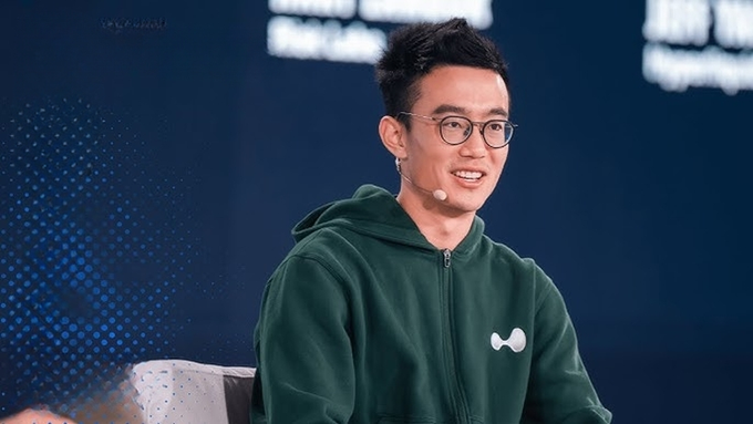 A man wearing glasses and a green hoodie with a bone logo sits smiling in what appears to be a studio or interview setting.