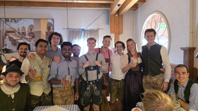A group of people dressed in traditional Bavarian attire, including lederhosen and dirndls, posing together and holding large beer mugs inside a wooden-beamed hall.
