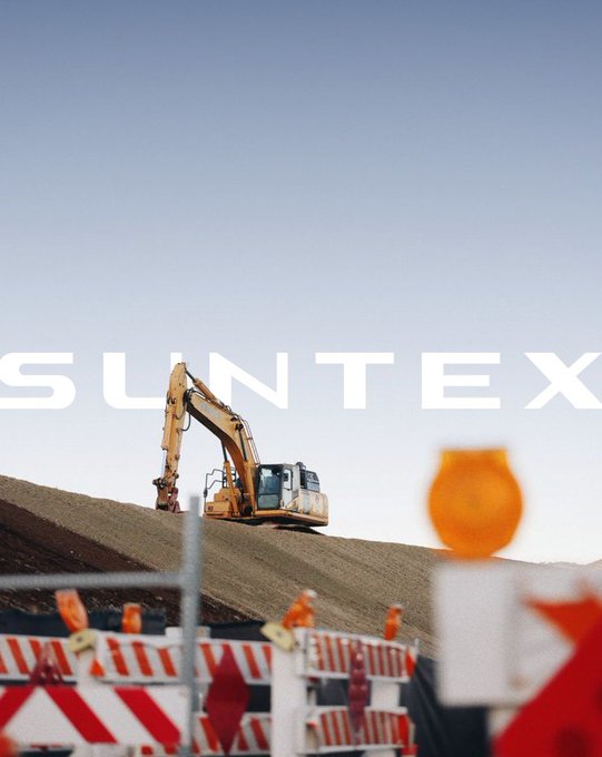 A construction scene featuring an excavator on a hill of dirt with the large white text 'SUNTEX' overlaid across the middle of the image, and a blurred orange warning light in the foreground.
