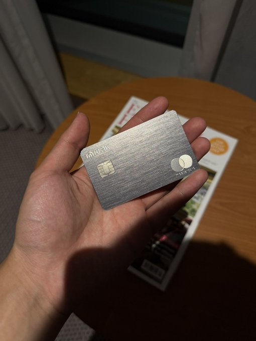 A hand holding a silver metallic credit card over a round wooden table with some magazines or brochures.