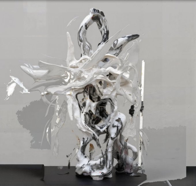 A glossy, abstract white sculpture with marbled black veining stands on a dark surface, its fluid, dripping forms creating a chaotic yet sculptural composition. The piece reflects the artist's statement about integrating AI with traditional media—its organic, hybrid texture blurring the line between digital generation and physical materiality, as if a 3D render has been pulled into tangible form.