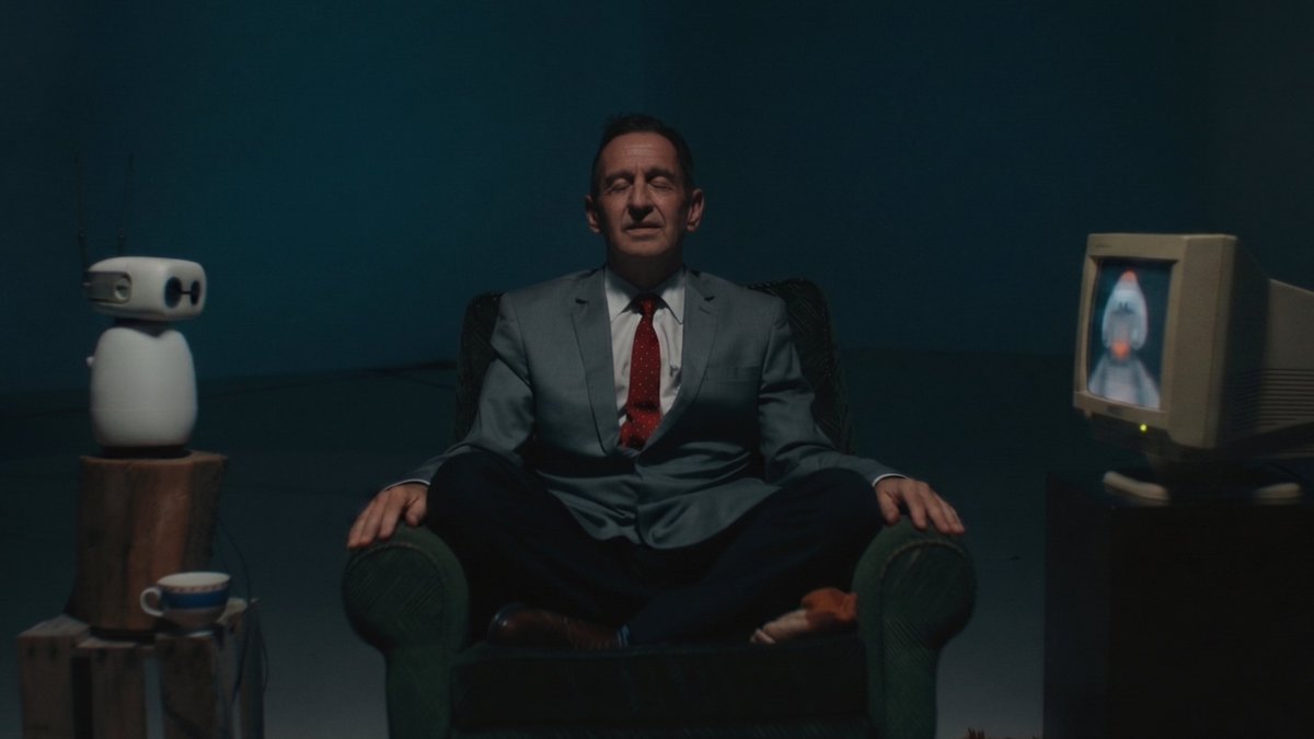 A man in a gray suit and red tie sits cross-legged in a green armchair with his eyes closed, flanked by a small white robot and an old CRT television displaying a strange image.