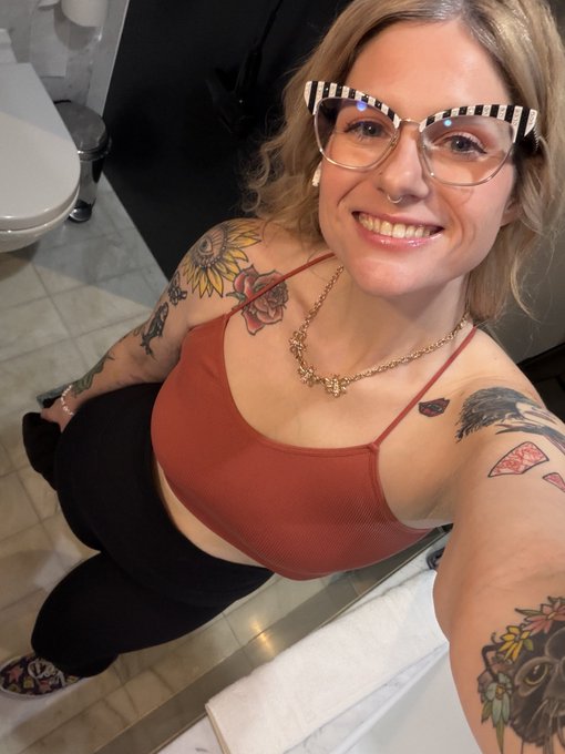 A selfie of a smiling woman with blonde hair, black and white striped glasses, a nose ring, and multiple tattoos, wearing a red tank top and gold necklace in what appears to be a bathroom.