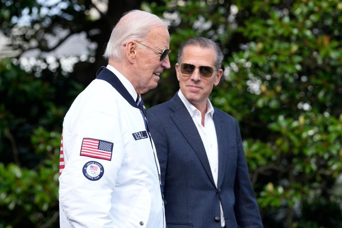 Two men wearing sunglasses stand outdoors with trees behind them; one wears a white NASA jacket with an American flag patch, while the other is dressed in a dark gray suit.