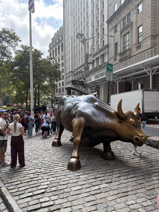 A photo of the iconic Charging Bull bronze statue on Wall Street in New York City, with tourists gathered around it on the cobblestone street.