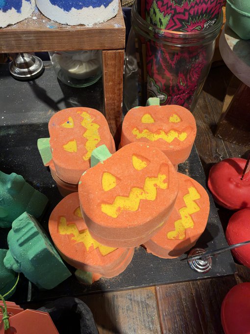 A collection of Halloween-themed pumpkin-shaped bath bombs with jack-o'-lantern faces stacked on a dark surface alongside other bath products.