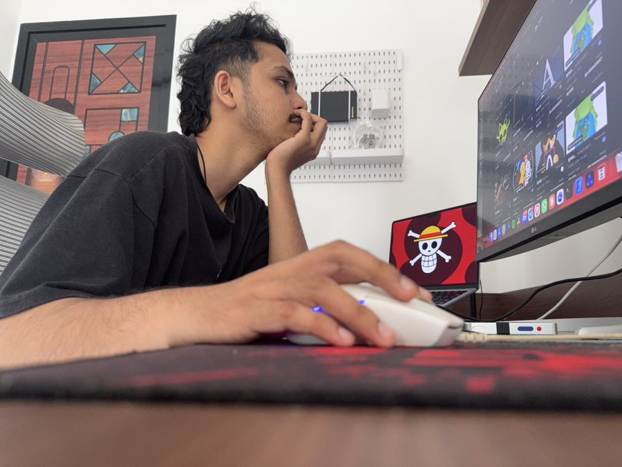 A young man with curly hair sits at a desk using a white computer mouse while looking at a monitor, with a laptop displaying a Straw Hat Pirates logo visible in the background.