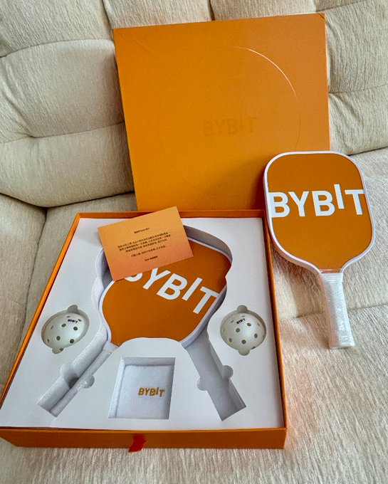 An unboxed orange gift set from 'BYBIT' containing branded pickleball paddles, balls, and a small card, displayed on a beige textured surface.