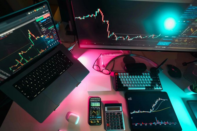 A trader's multi-screen workstation displays candlestick charts and market data across a laptop, monitor, and tablet, illuminated by pink and teal neon lighting. The setup, complete with keyboard, calculator, and smartphone, captures the tense, data-driven atmosphere as Bitcoin and Ether traders brace for volatile swings ahead of today's U.S. CPI inflation release.