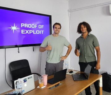Two men standing in an office space, both giving thumbs up, with a monitor behind them displaying 'PROOF OF EXPLOIT' on a purple background, and a desk with laptops and a first aid kit in the foreground.