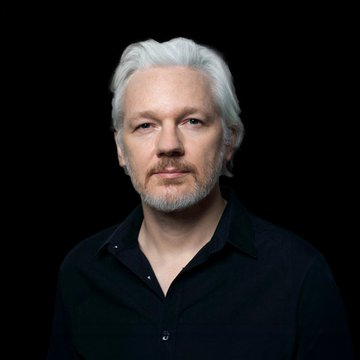 Portrait of Julian Assange, the WikiLeaks founder, with white hair and beard, wearing a black button-up shirt against a dark background.