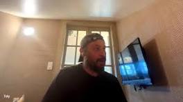 A webcam-style view of a bearded man wearing a beanie in a dimly lit room, with a computer monitor displaying a desktop visible in the background.