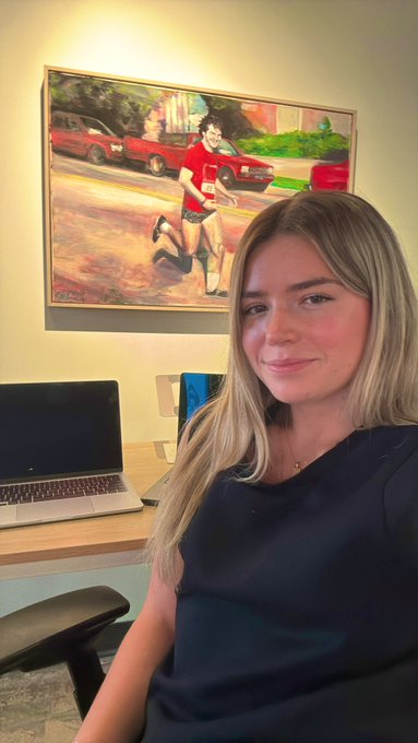 The image features a woman sitting down with her laptop open before her. She appears to be smiling for the camera while working or browsing the internet. The background of the photo shows an art piece, possibly a painting, which adds an artistic touch to the scene. The text in the image reads "Started my first big girl job yesterday! Excited to join @BitcoinMagazine as an Associate Producer working on guest programming for BMTV - Bitcoin Magazine TV show!"