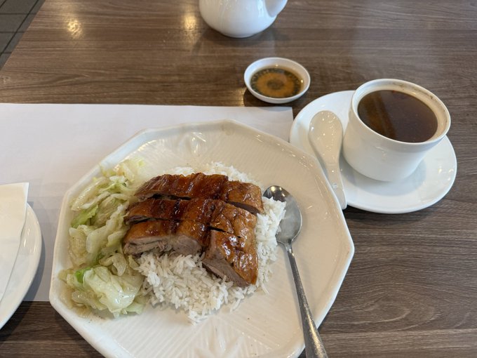 A meal consisting of sliced roasted duck or pork over white rice with cabbage on an octagonal plate, accompanied by a cup of dark coffee/tea and a small dish of dipping sauce on a wooden table.
