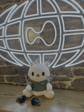 A cute white plush toy cat wearing a green vest sits in front of a white neon sign depicting a globe with a carved-out piece, on a wooden floor near a brick wall.