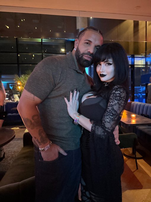 A photograph of a bearded man with tattoos on his arm wearing sunglasses on his head, posing alongside a woman with long black hair, pale makeup, and dark lipstick dressed in a black lace outfit.
