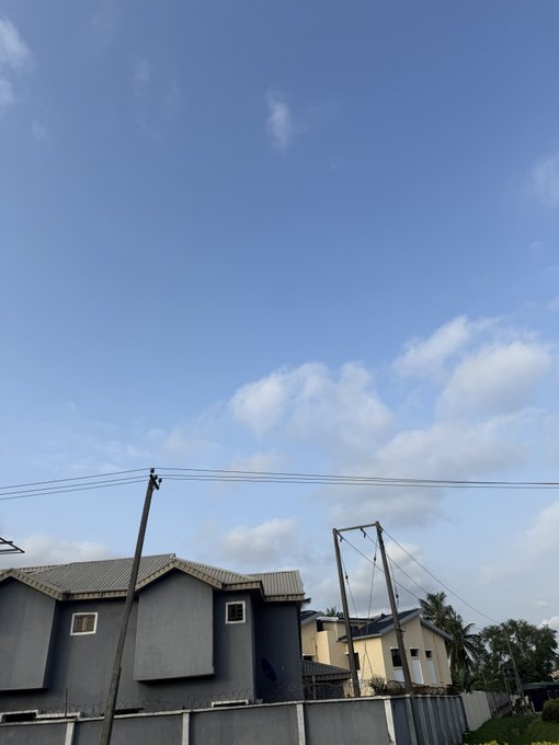 A residential neighborhood under a bright blue sky, with gray houses, utility poles, power lines, trees, and a palm tree visible.