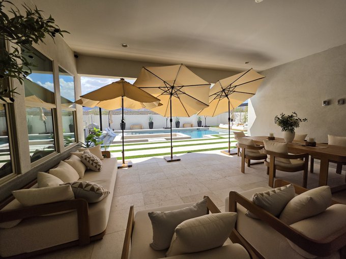 A luxurious outdoor patio area featuring cream-colored sofas with pillows, three large tan umbrellas, a swimming pool, and a dining set, all set against a sunny sky.