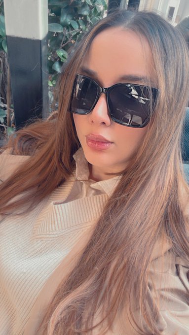 A selfie of a woman with long brown hair wearing large black sunglasses and a beige top.