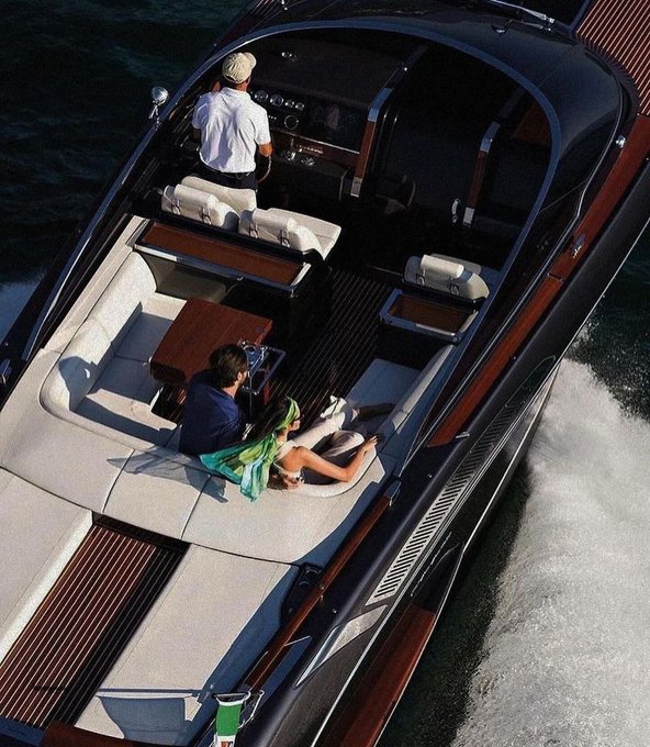 An aerial view shows a luxury wooden speedboat cruising through the water, with a captain at the helm and two passengers relaxing in the seating area below.