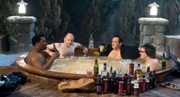 Four men relaxing in a bubbling wooden hot tub at night, surrounded by numerous beer bottles and cans on the edge of the tub, set in a snowy, lantern-lit environment.
