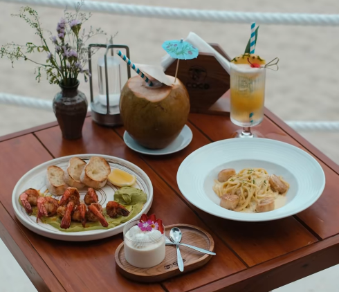 A wooden table by a beach rope barrier displaying a spread of food including lobster/avocado appetizer, bread, pasta, a coconut drink with blue paper umbrella, a yellow cocktail, and a small dessert, with a vase of wildflowers.