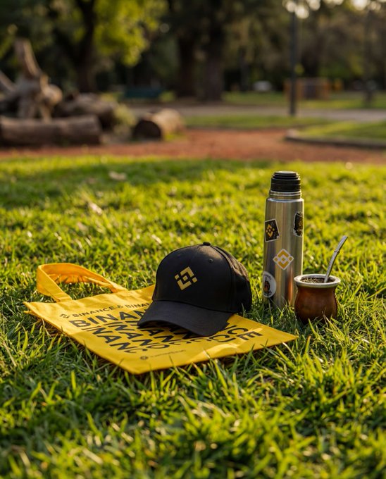 A black cap with a Binance logo resting on a yellow Binance drawstring bag on grass, next to a thermos and a mate gourd with straw.