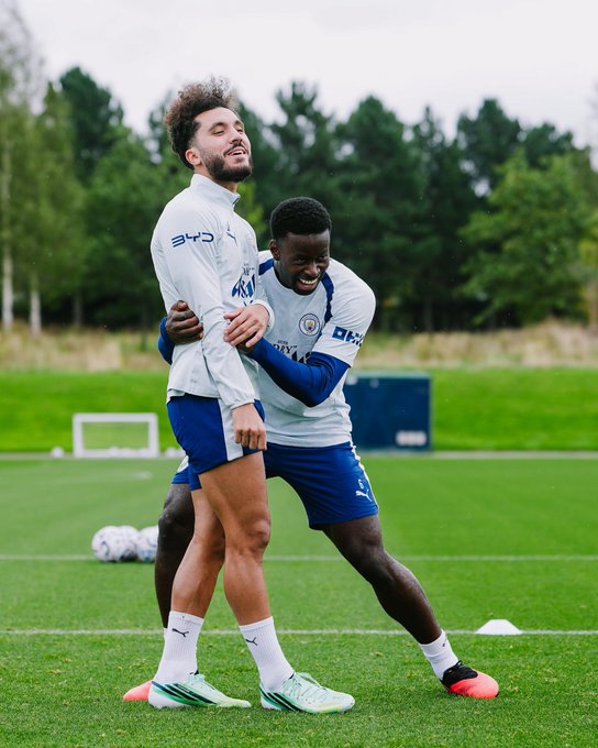 Two Manchester City football players in training gear joke around on a practice pitch, with one grabbing the other from behind while both smile and laugh.
