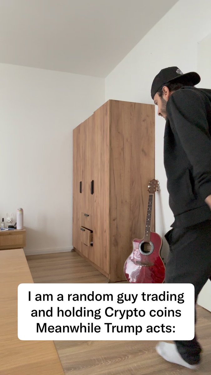 A man in dark clothing and a black cap moves energetically in a room with a wooden wardrobe and red acoustic guitar; the caption reads "I am a random guy trading and holding Crypto coins Meanwhile Trump acts:".
