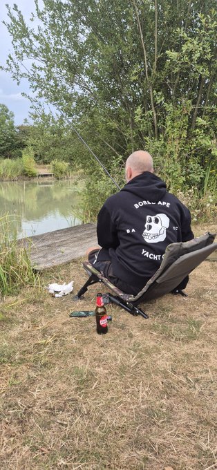 A bald man wearing a black hoodie with a skull design and the text 'GORL AFE' and 'YACHTS' sits in a camping chair fishing by a lake, with a bottle of soda on the ground beside him.