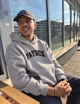 A man wearing a gray 'PAYBOX' hoodie and a black baseball cap sitting on a wooden bench and smiling at the camera.