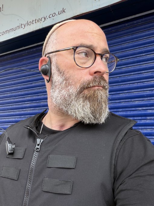 A bald man with a gray beard wearing a beige kippah, round glasses, and an earpiece, dressed in a black vest over a black shirt, standing in front of a blue shutter with 'communitylottery.co.uk' visible above.