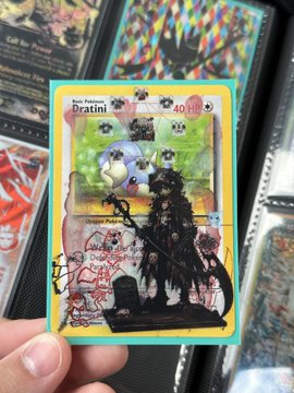 The image features a hand holding up a Pokemon card with a unique design featuring a black figure of a dragon, surrounded by various other Pokemon cards. The person's hand is positioned over the card, drawing attention to its distinct appearance. The background includes more Pokemon cards and a description that reads "focus on what is in the image and how it relates to the post". This suggests that the image might be related to a discussion or analysis of the Pokemon card design, possibly involving the concept of digital art or the value of such items as collectibles.
