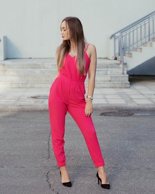 A fashion photo of a young woman with long brown hair wearing a hot pink jumpsuit and black high heels, posing on a paved area near concrete steps.