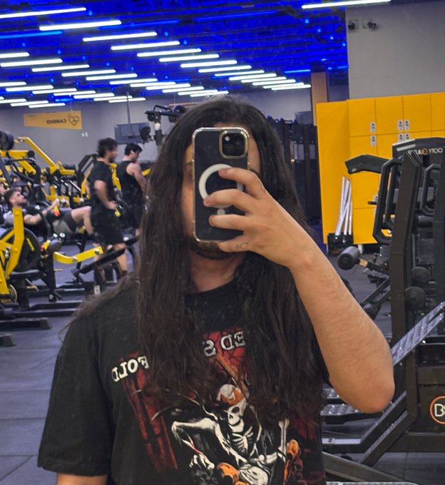 A mirror selfie of a long-haired man taking a photo in a gym with blue ceiling lights and yellow weight machines in the background, wearing a black band t-shirt.