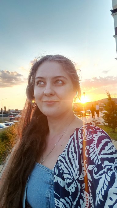 A young woman with long brown hair wearing a blue patterned top, taking a selfie during sunset.